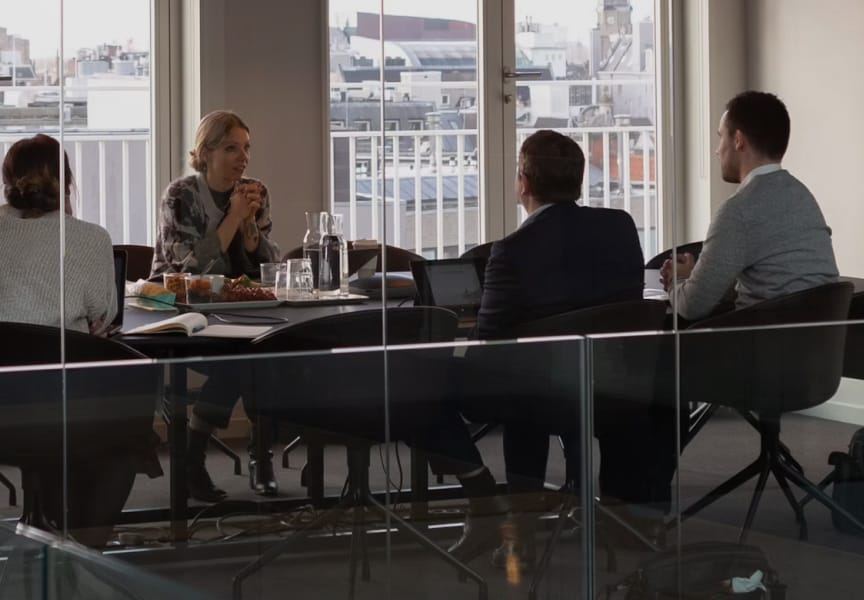 Business meeting in a modern office setting with participants discussing around a table, featuring views of the city skyline through windows.
