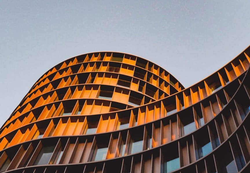 Curved architectural building facade with a series of orange and brown panels reflecting light against a clear sky.