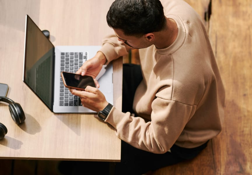 Person using a laptop and smartphone while seated at a wooden table, with headphones visible next to the laptop.