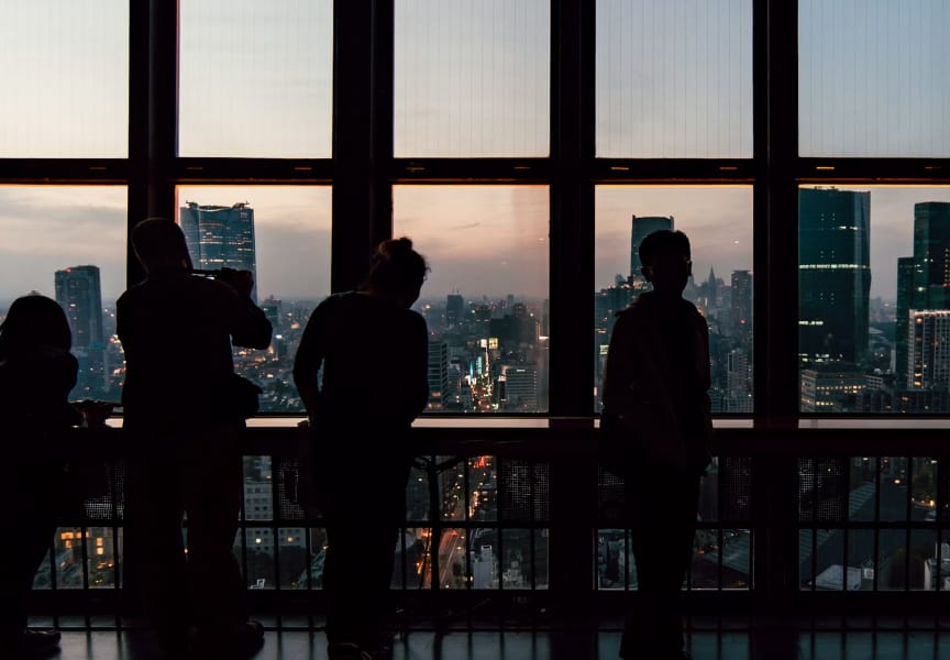 Silhouetted figures observing a city skyline at dusk, with tall buildings and a sunset in the background.