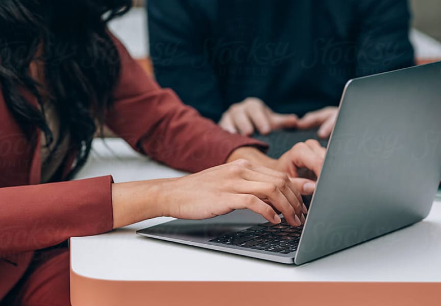 A close-up of hands typing on a laptop, with another person in the background working on a different device at a desk.