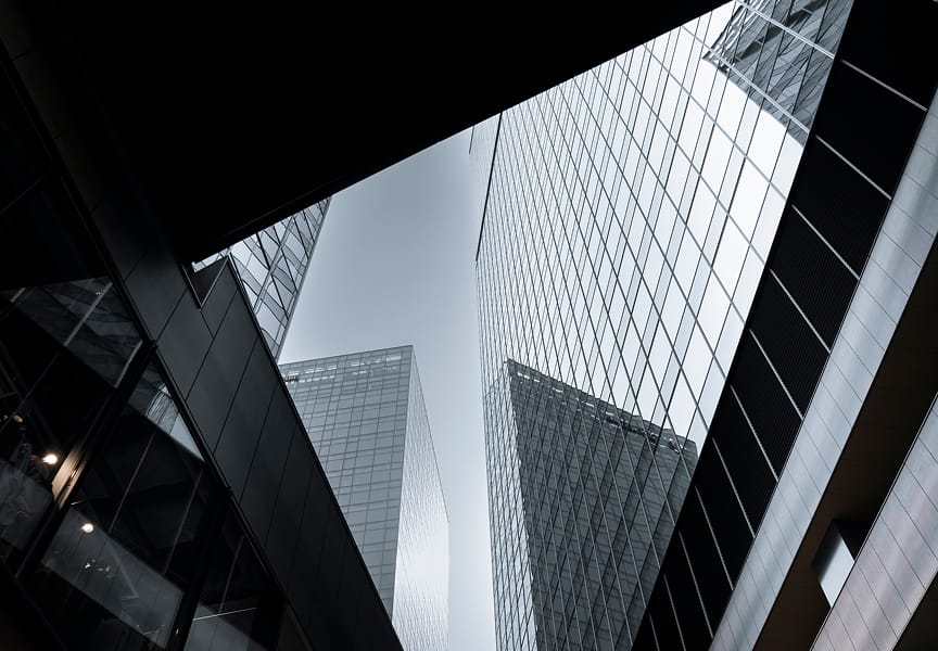 View of towering glass skyscrapers from a narrow angle, showcasing reflective surfaces and geometric lines against a bright sky.