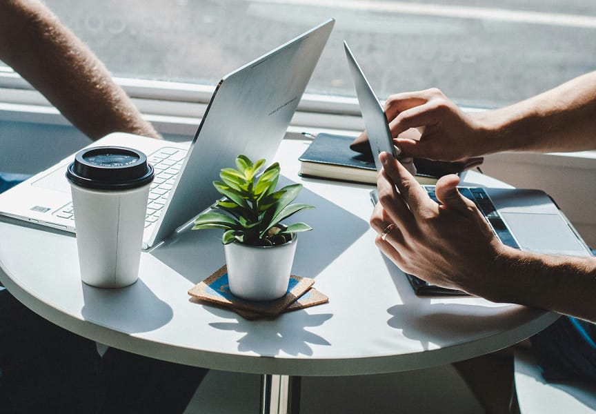 Laptop and tablet on a table with a coffee cup and a small potted plant, with hands visible working on the devices.