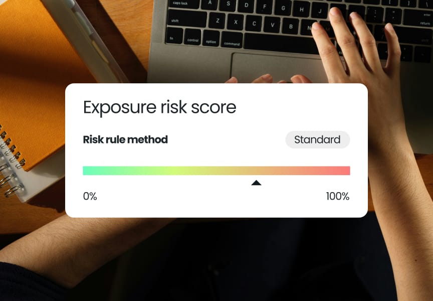 Exposure risk score display with a gradient scale from 0% to 100%, shown above hands on a laptop keyboard and a notebook nearby.