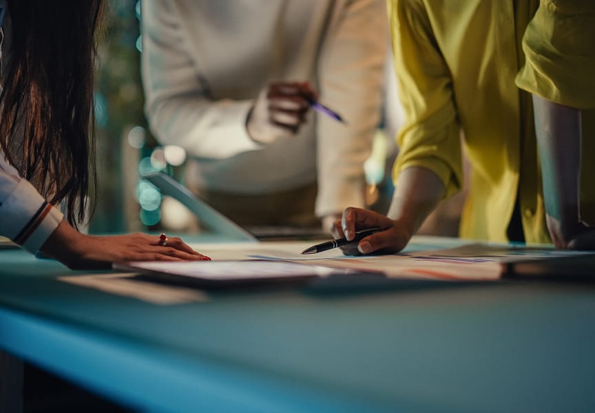 Hands of diverse individuals discussing documents and making notes on a table, with soft lighting in a collaborative workspace.