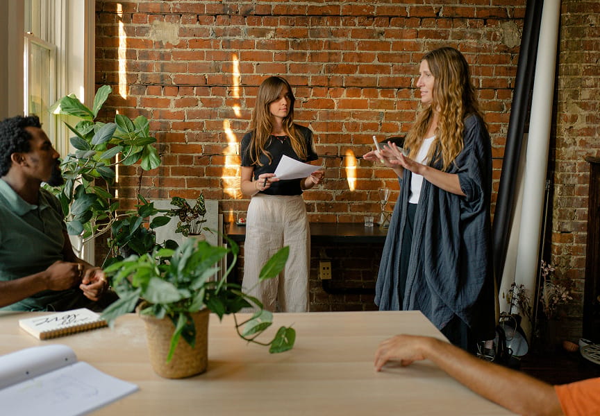 Two individuals are engaged in conversation in a cozy, brick-walled setting, with plants and a table in view. One person holds a paper.