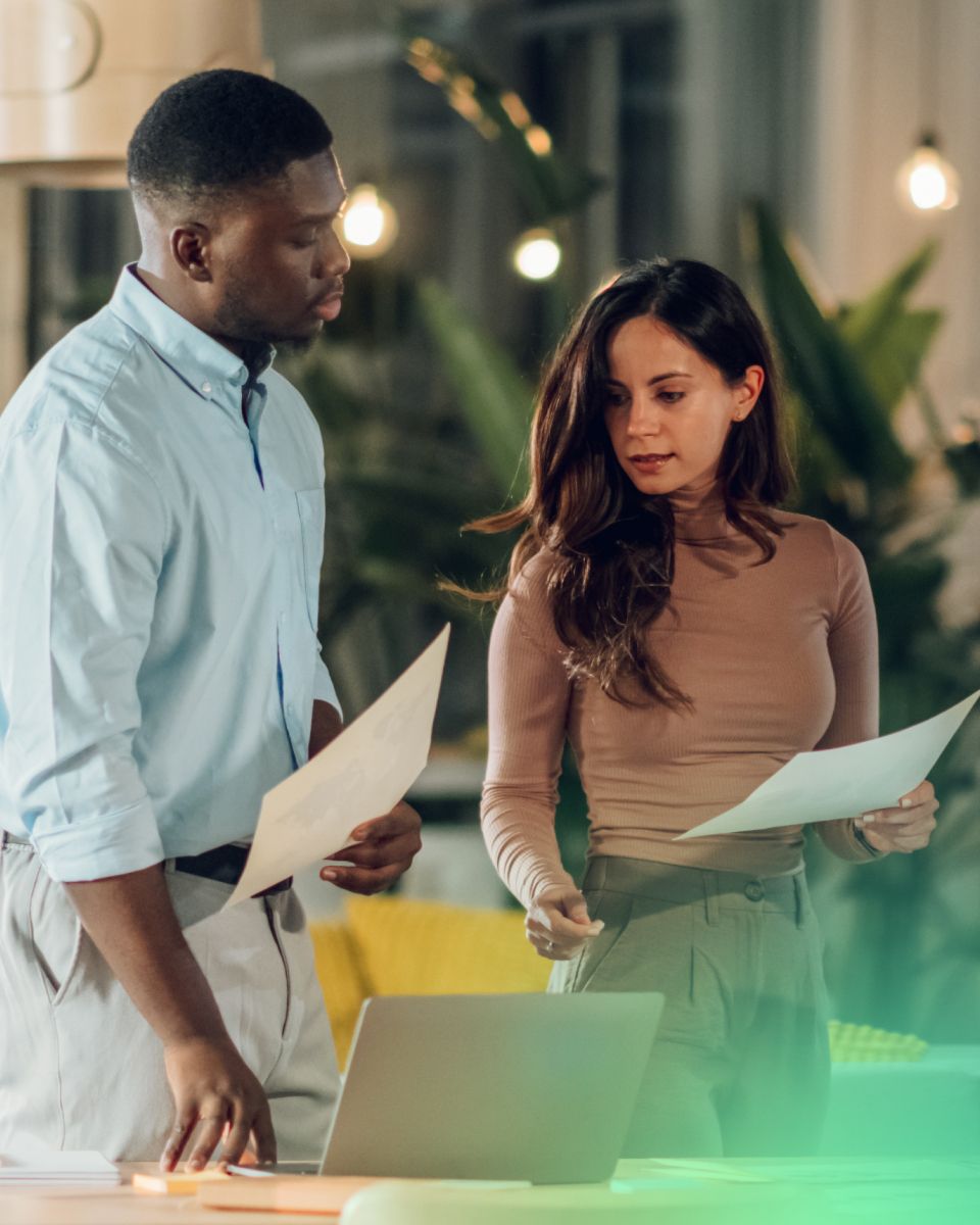 Two individuals discuss documents while standing in a modern workspace, surrounded by plants and soft lighting, with a laptop visible on the table.