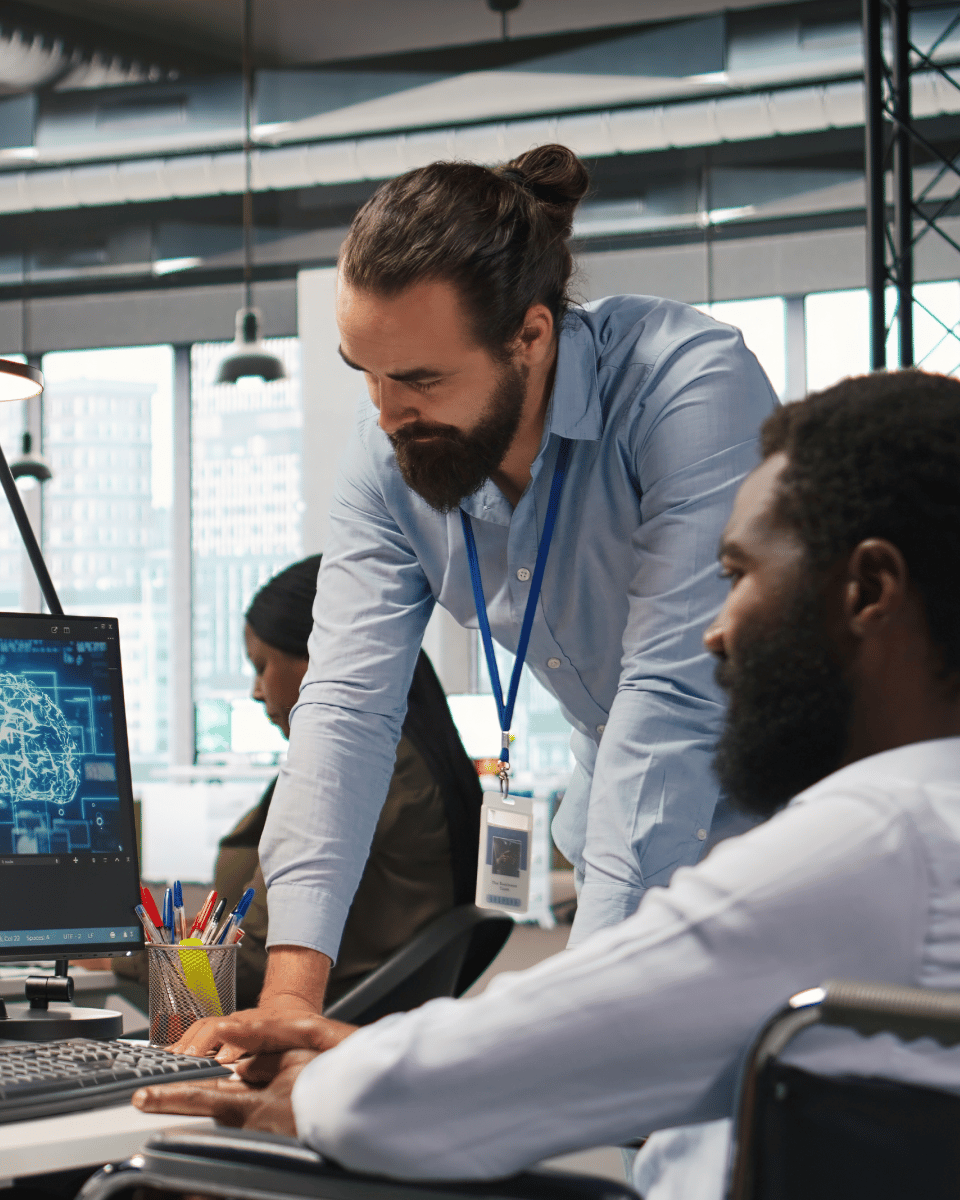 Two individuals collaborating in an office environment, with one analyzing data on a computer screen while the other observes and provides input.
