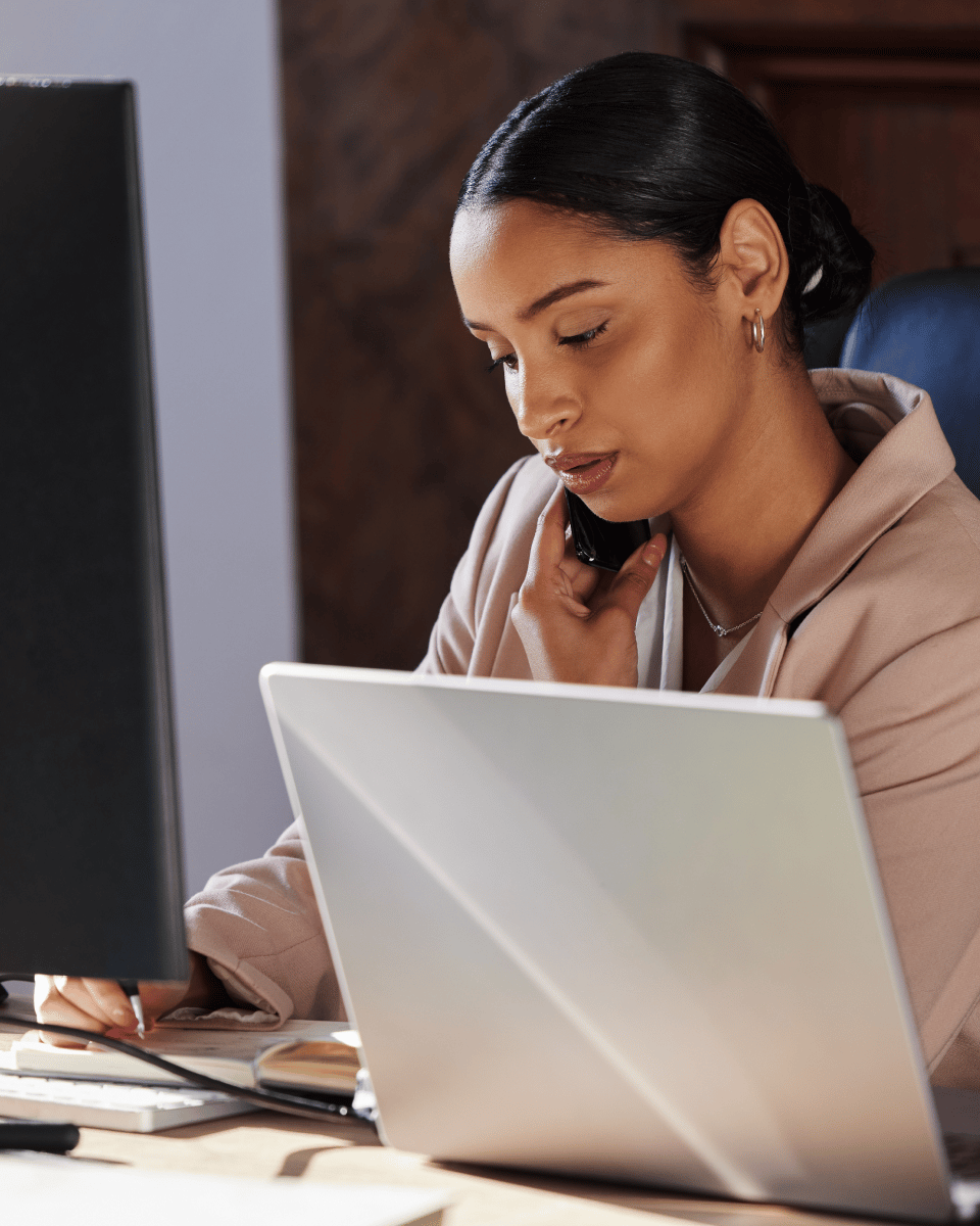 A person engaged in a phone conversation while taking notes, seated at a desk with two laptops.