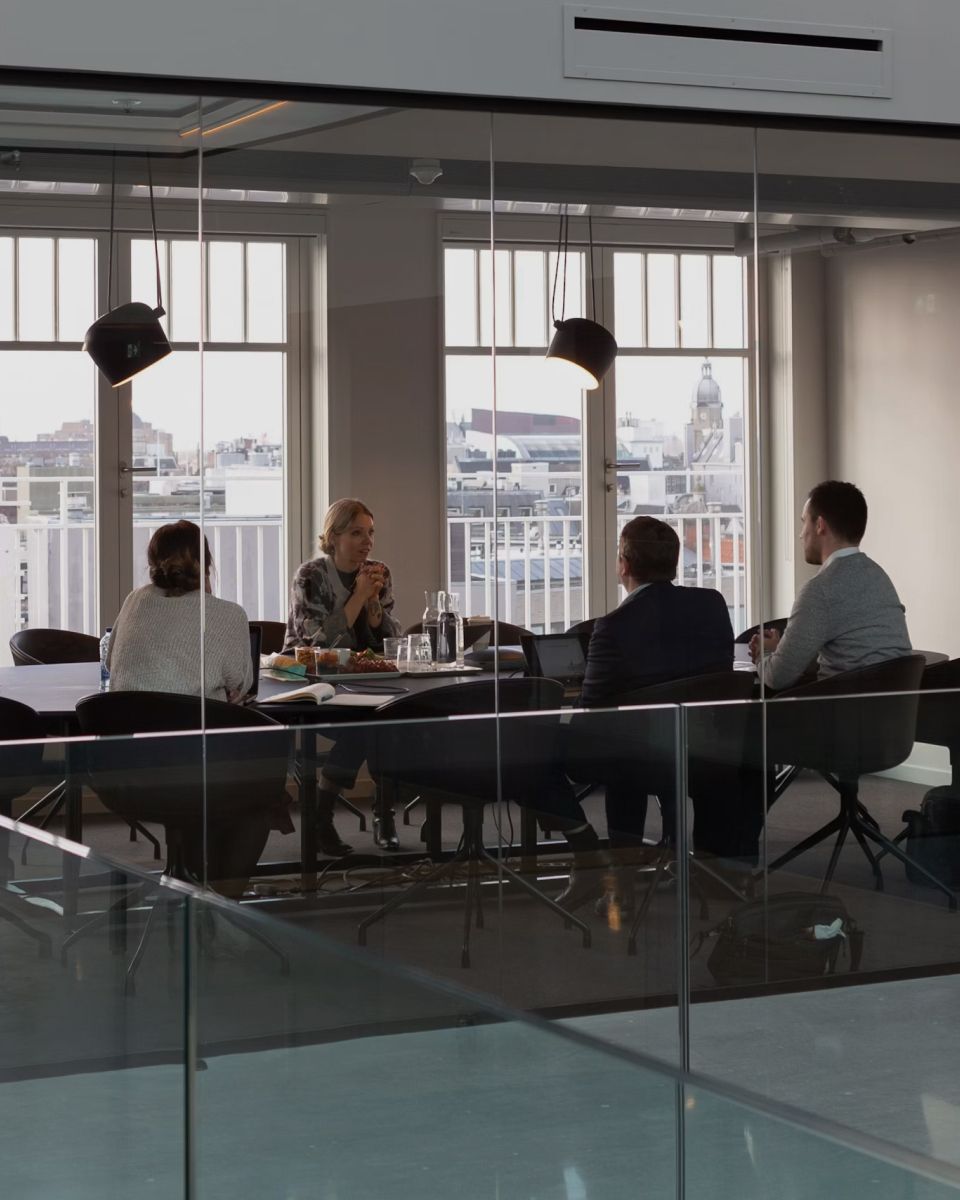 A meeting taking place in a modern office with large windows, showcasing a city view and a group discussing around a table.