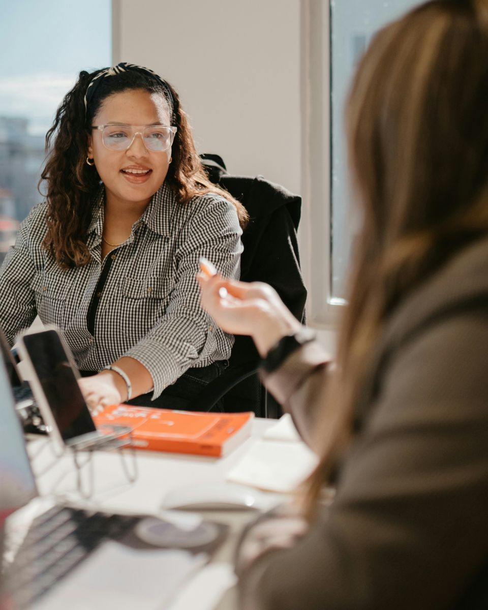 Two individuals engaged in a discussion at a modern workspace, with laptops and a notebook on the table. Natural light from a window.