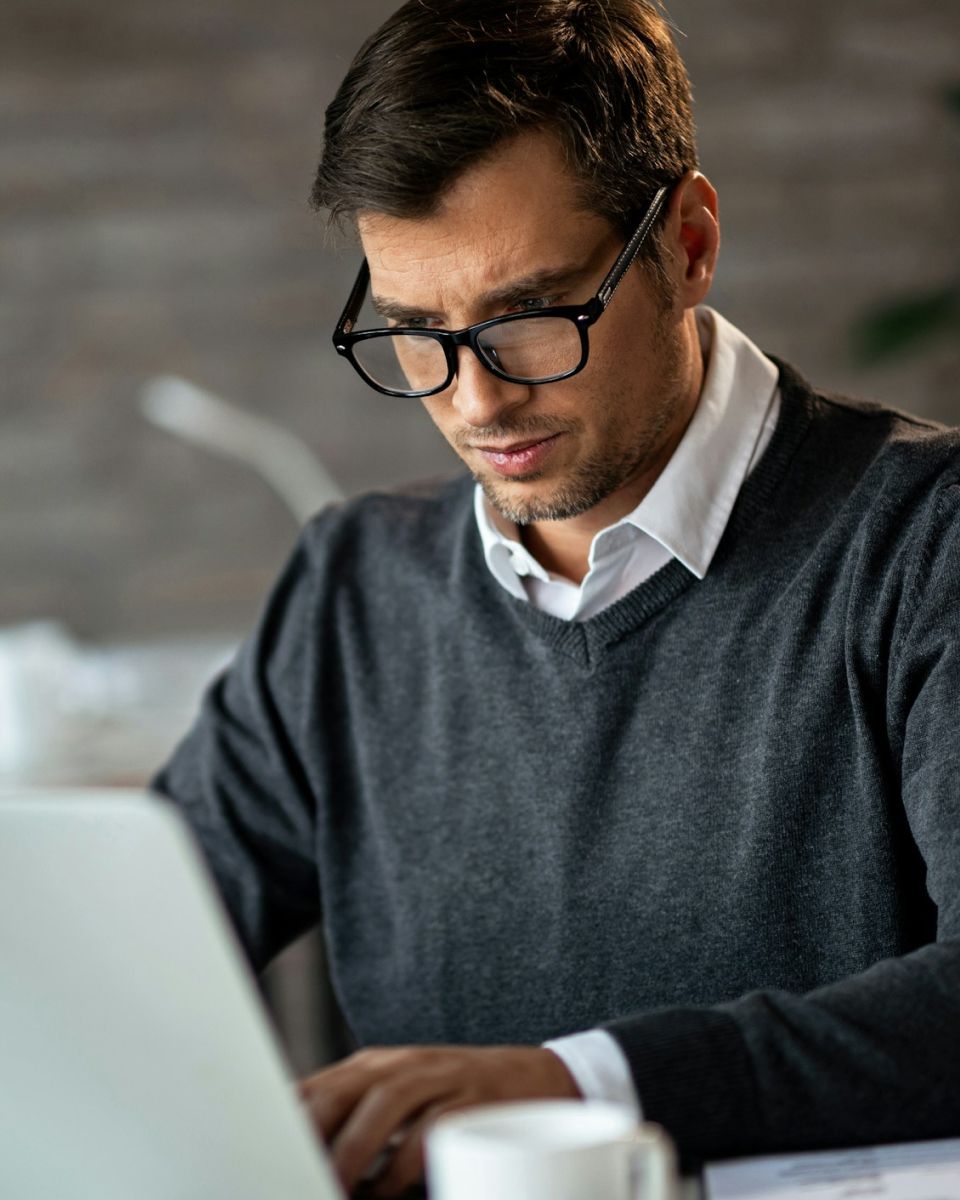 Professional headshot of a person working on a laptop, wearing glasses and a sweater in a modern office setting.