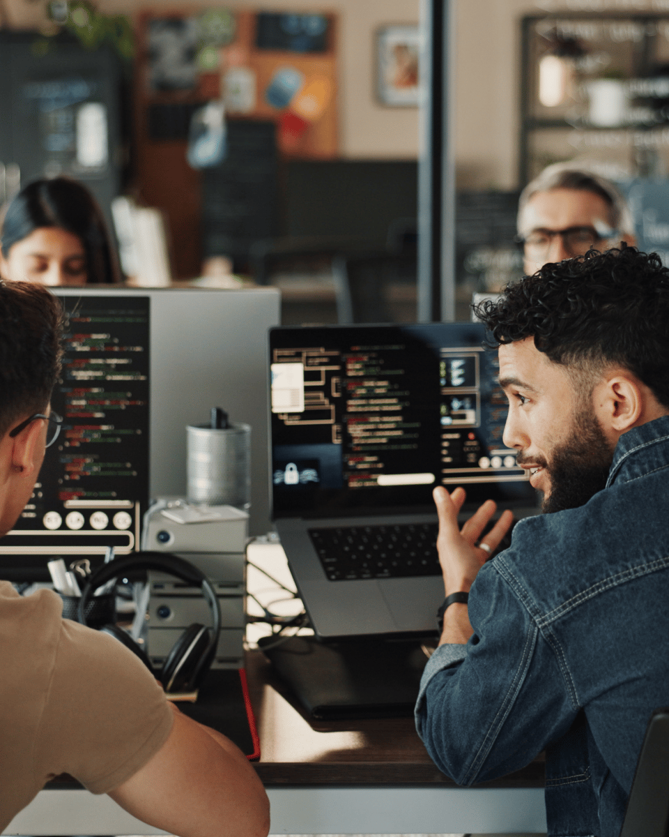 A diverse group of people working collaboratively in a modern office, with one person gesturing while discussing on a laptop.