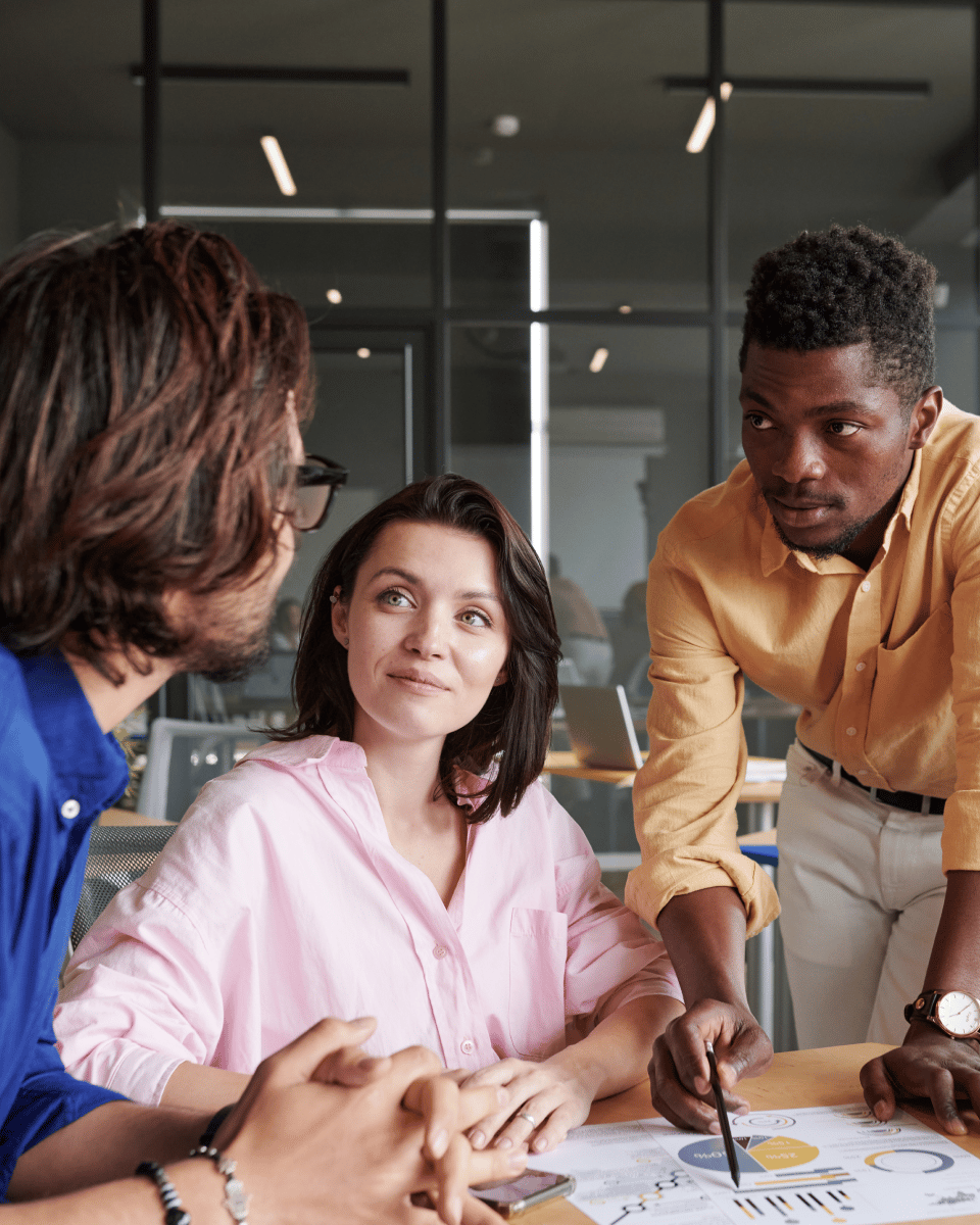 Three individuals engaged in a discussion around a table with charts and graphs, set in a modern office environment.