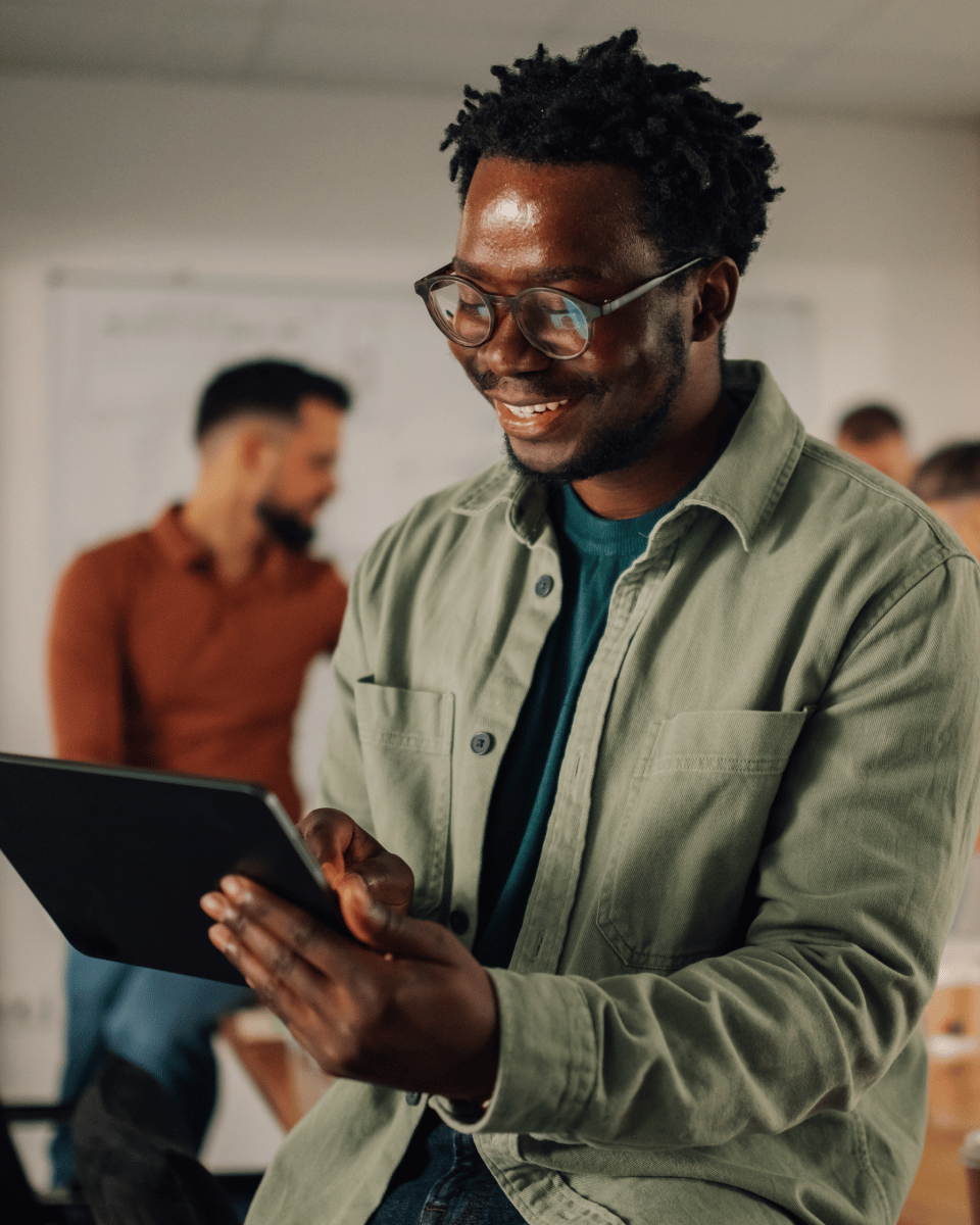 Smiling individual using a tablet in a modern office setting with colleagues in the background engaged in conversation.