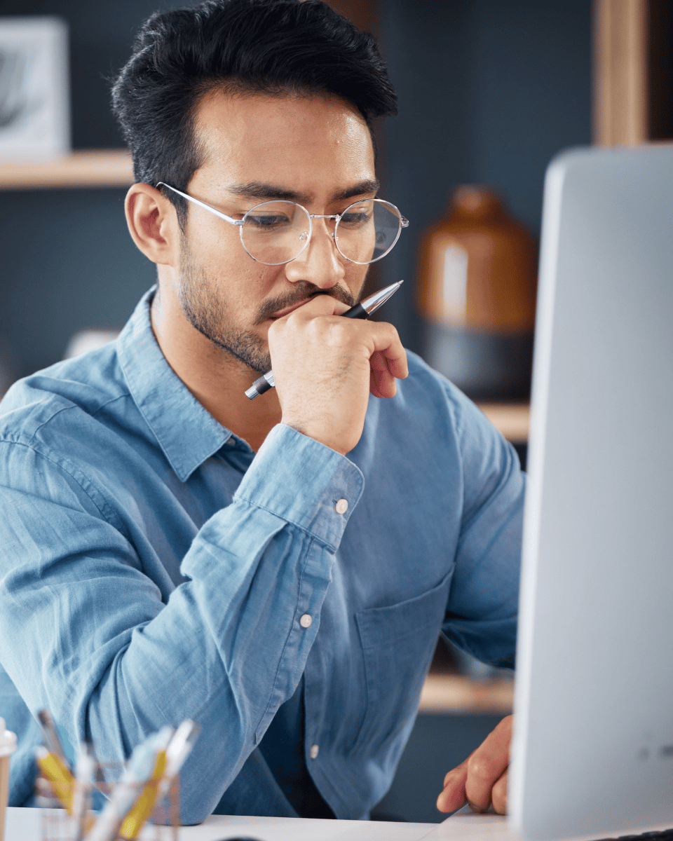 A focused individual working at a desk, holding a pen and looking thoughtfully at a computer screen, surrounded by a cozy office environment.