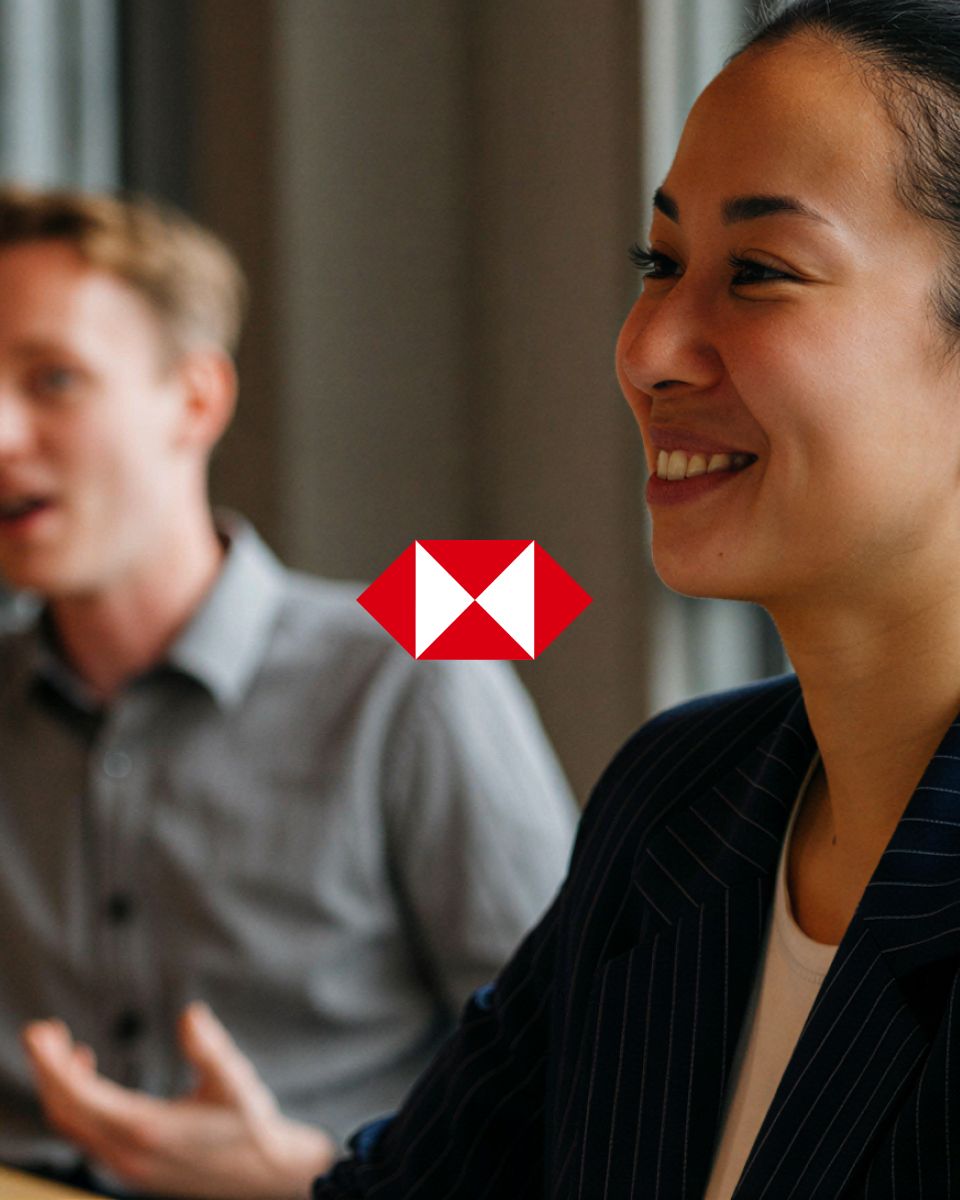 Smiling person in a tailored suit at a meeting, with a blurred figure speaking in the background and an HSBC logo in the foreground.